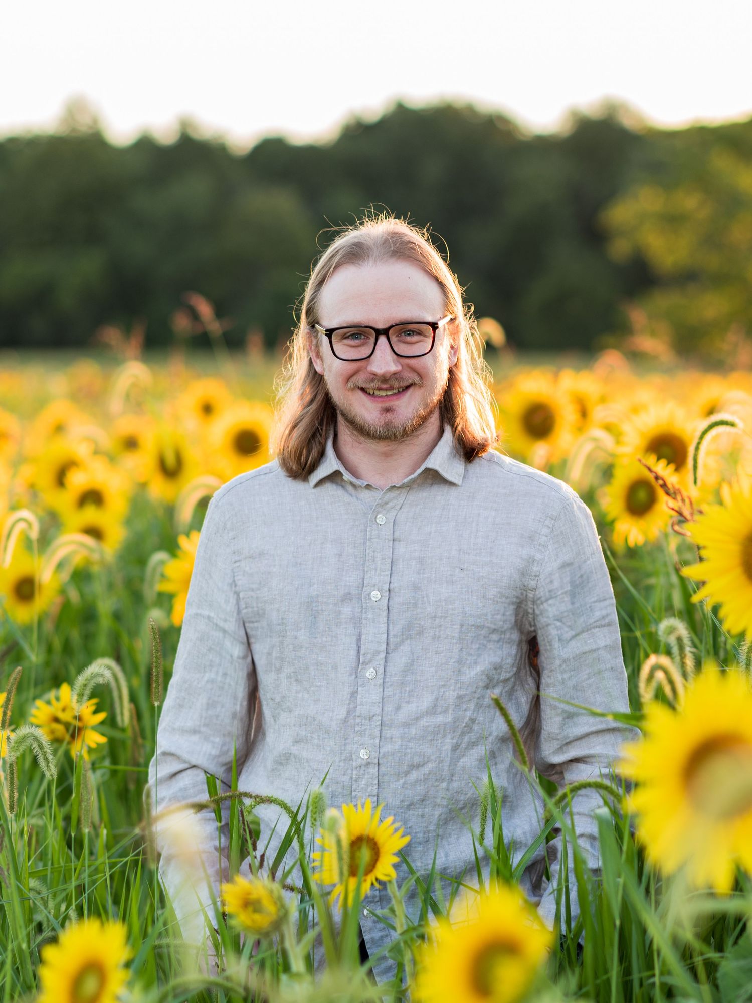 Person wearing glasses and linen shirt poses in bright sunflower field during golden hour.