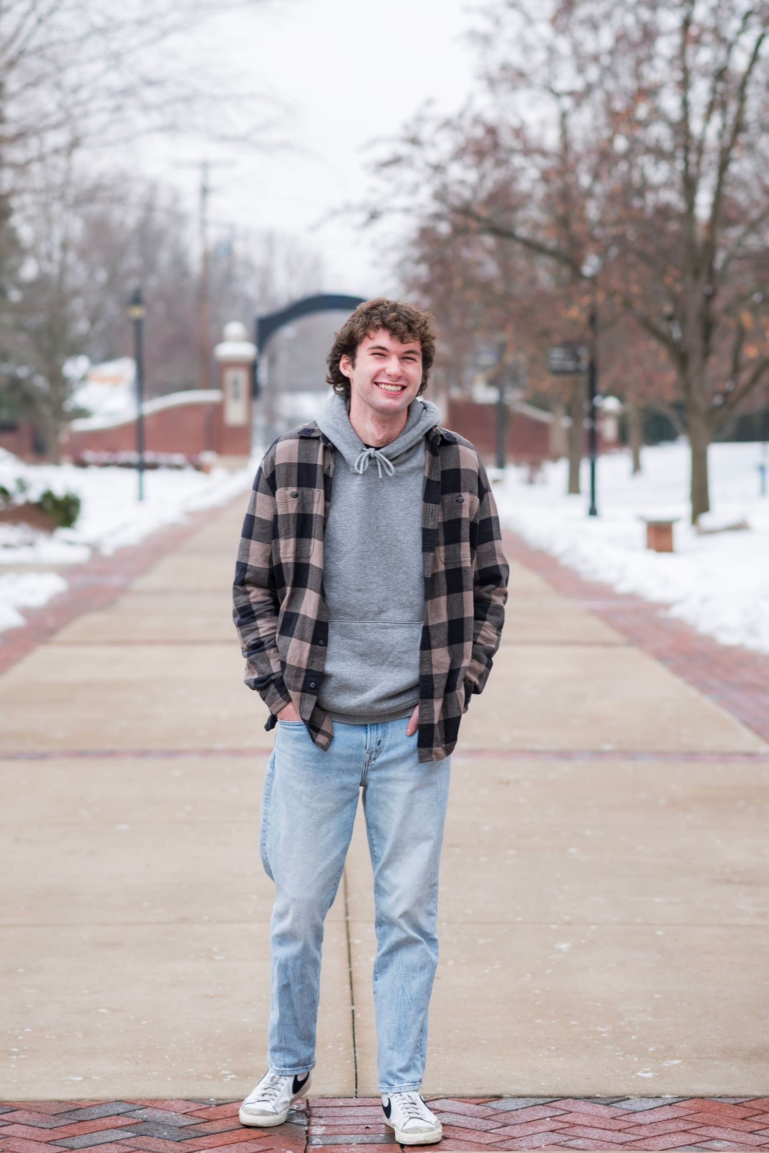 A casual winter scene of someone wearing a plaid flannel shirt and grey hoodie standing on a snowy campus walkway.