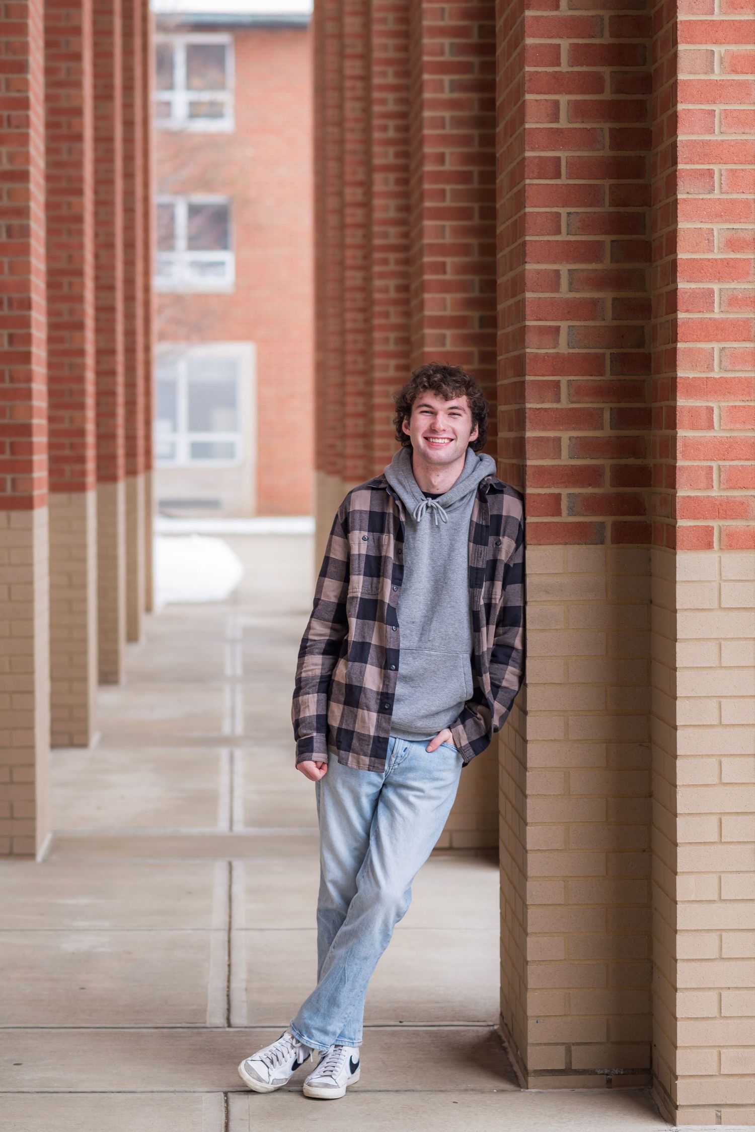 Person in plaid shirt and jeans poses casually against brick colonnade.