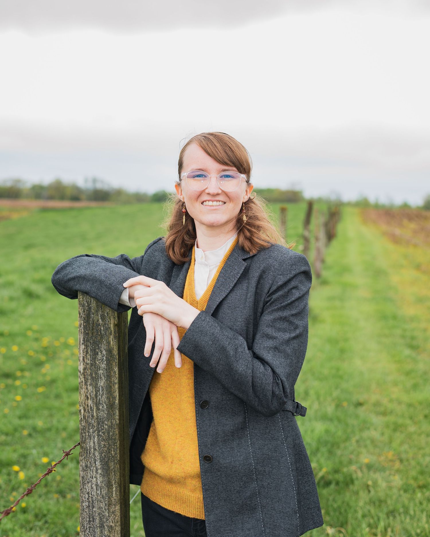 Someone in a yellow sweater and blazer leans against a rustic wooden fence in a countryside setting.