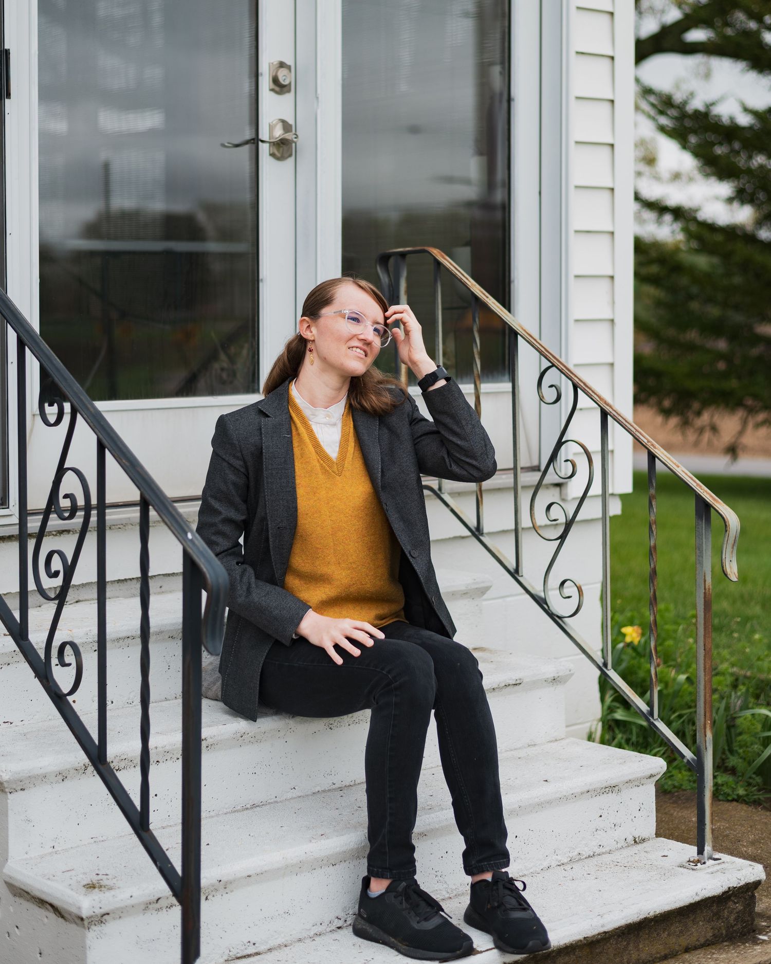 Someone in a mustard sweater and blazer sits casually on white porch steps with decorative railings.