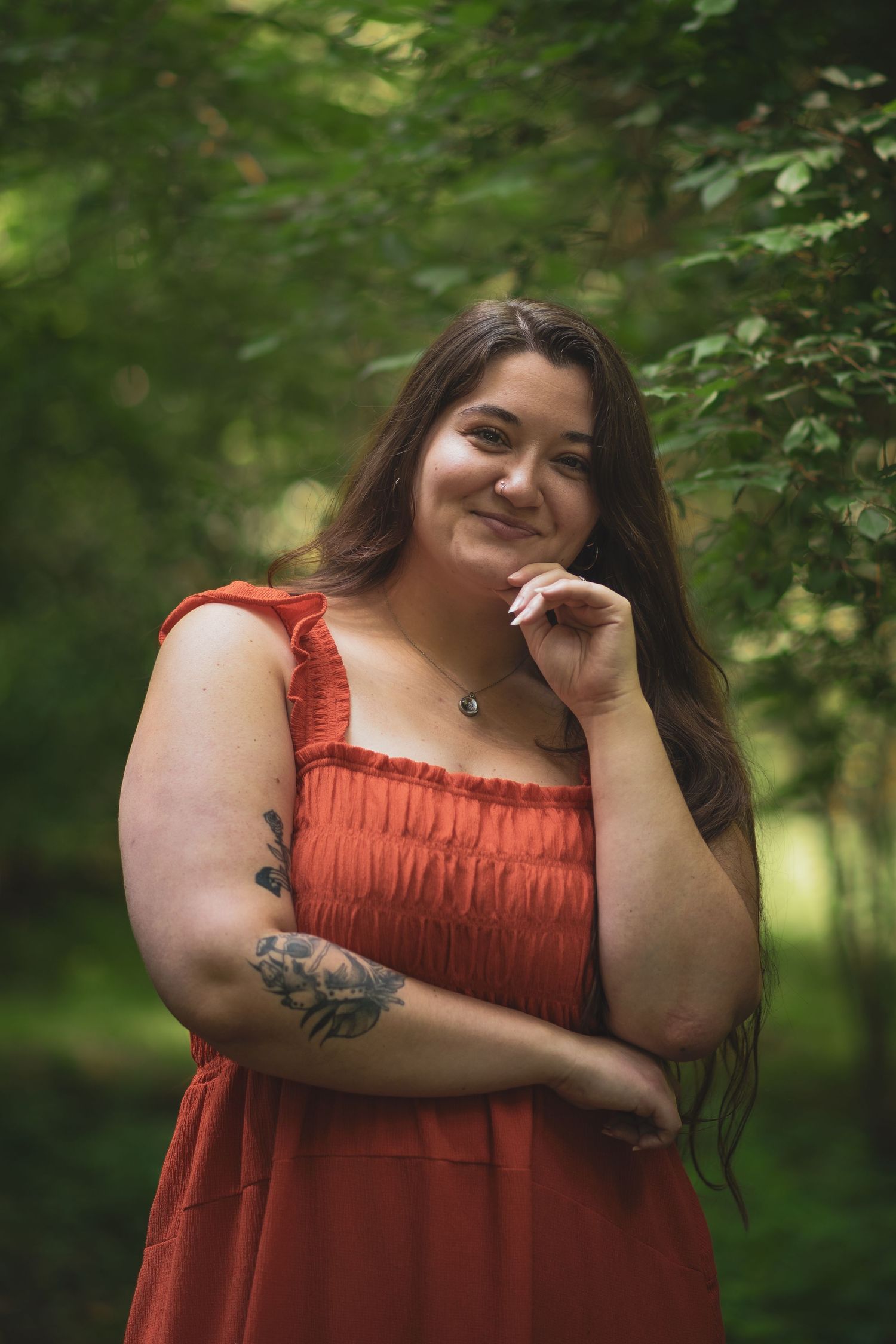 A person in a coral dress with tattoos poses playfully in a wooded area.
