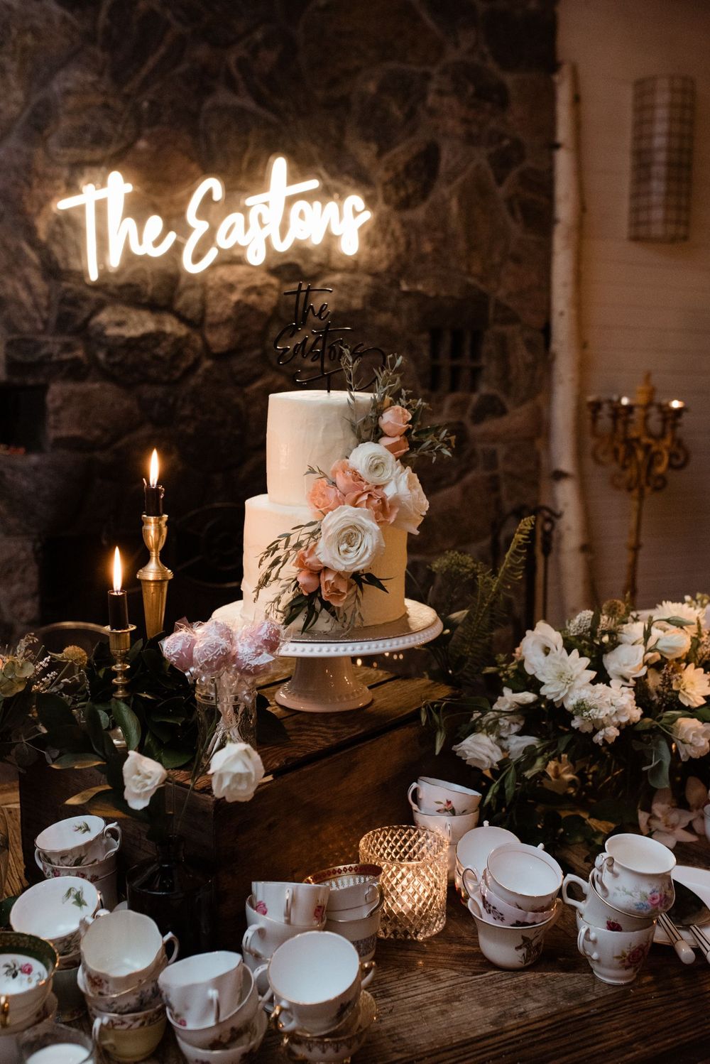 Elegant wedding cake display with neon sign reading 'The Eastons' illuminated on stone wall with vintage teacups and candles.
