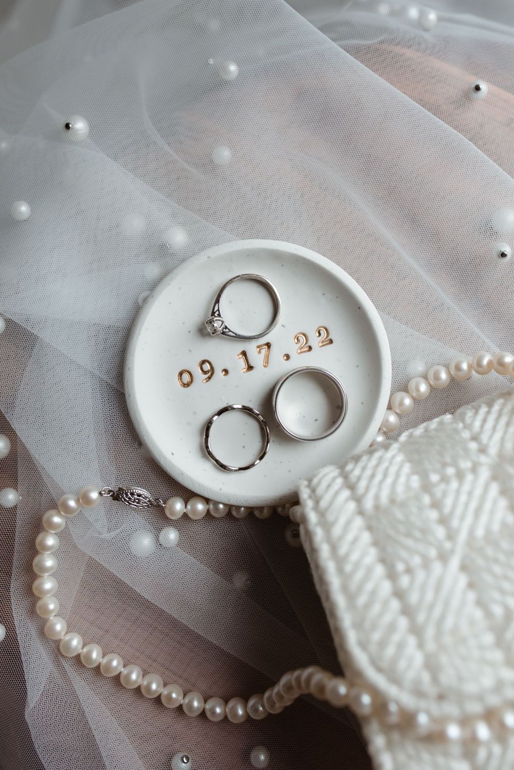 Wedding rings displayed on white ceramic ring dish with delicate pearls and tulle veil arrangements captured in detail.