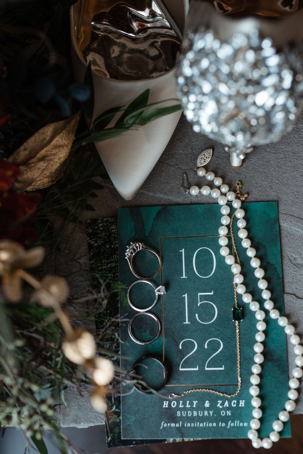 Wedding details arranged on dark surface showing rings, pearl necklace, and teal save-the-date card with date 10/15/22.