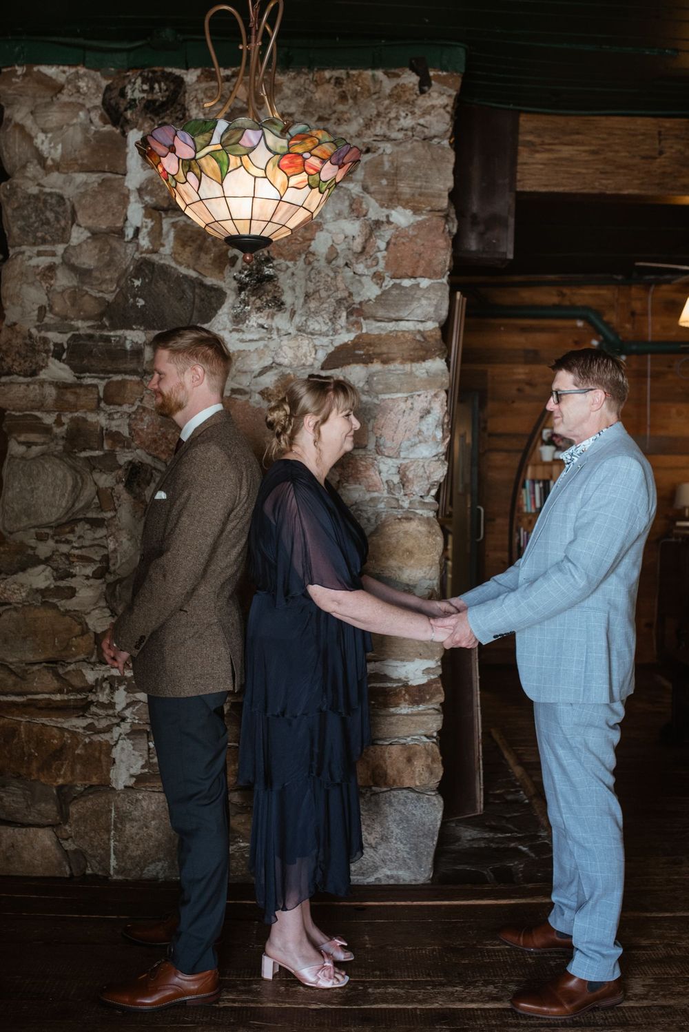 Three people stand in line against a rustic stone wall with a decorative Tiffany-style pendant lamp hanging above.