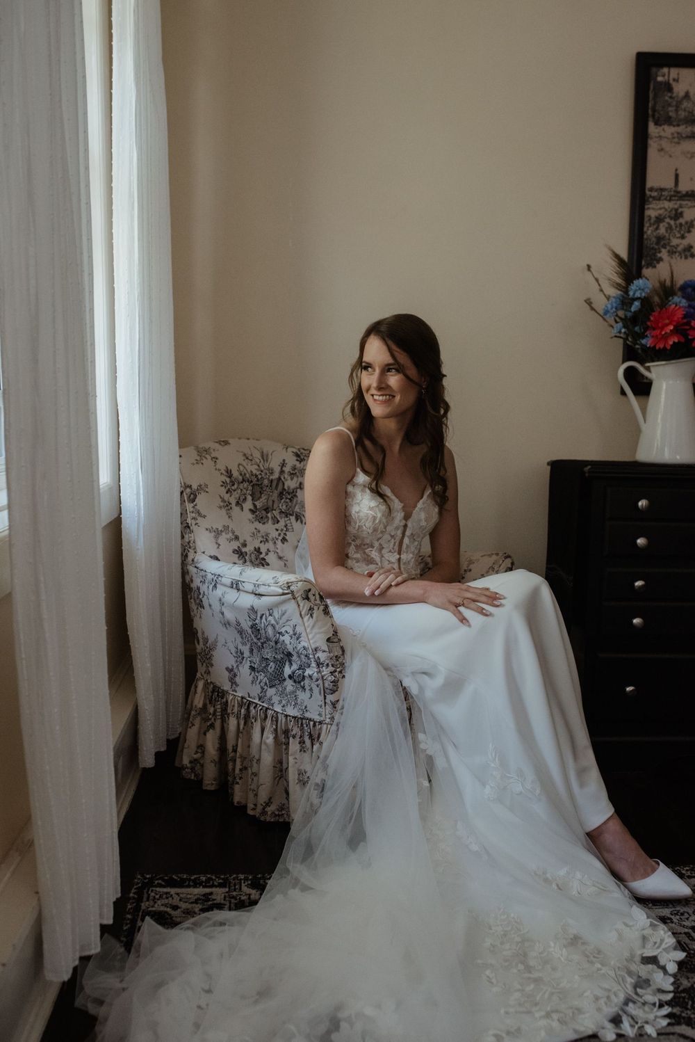 A bride in a flowing white gown with delicate lace detail sits elegantly on a patterned armchair near a window.
