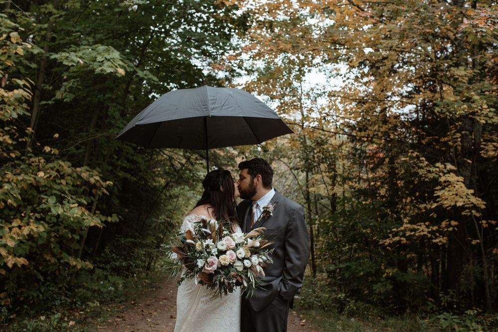 A couple shares a romantic moment under a black umbrella on a tree-lined path during an autumn wedding.