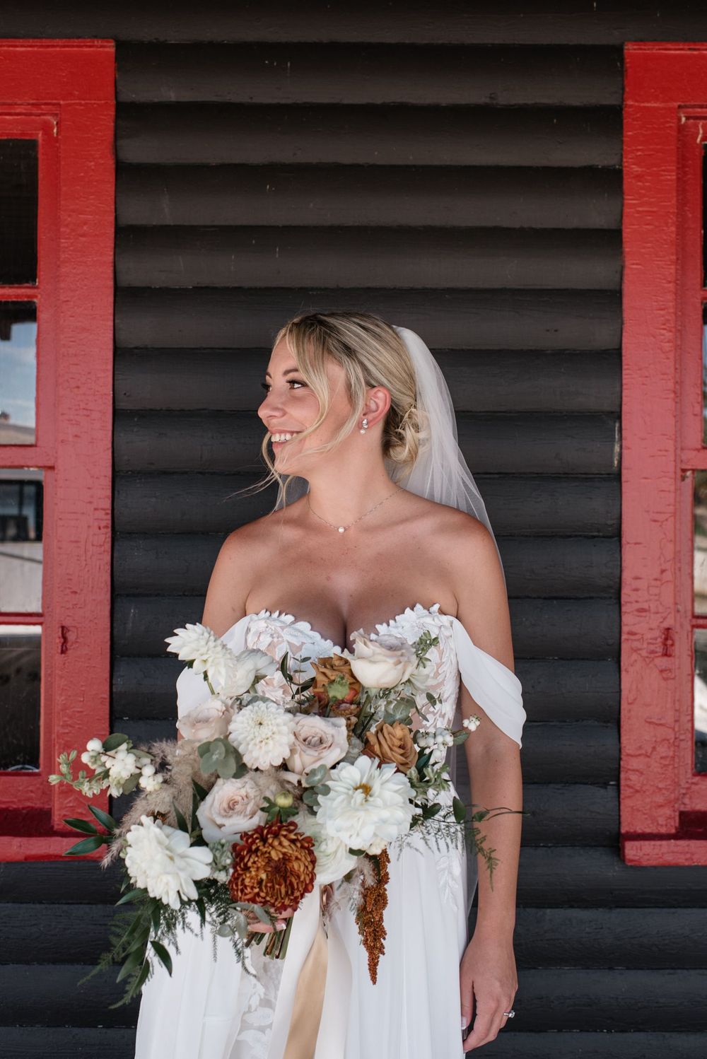 A bride in a white off-shoulder wedding dress holds a stunning bouquet of white and bronze flowers against a dark wall with red trim.