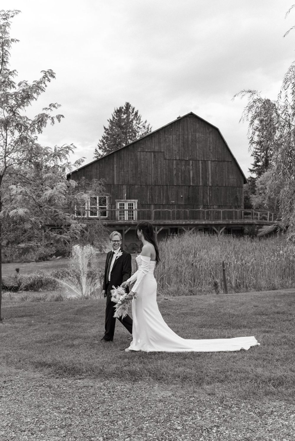 A gorgeous black and white wedding photo taken in front of a rustic wooden barn with a long flowing bridal dress.