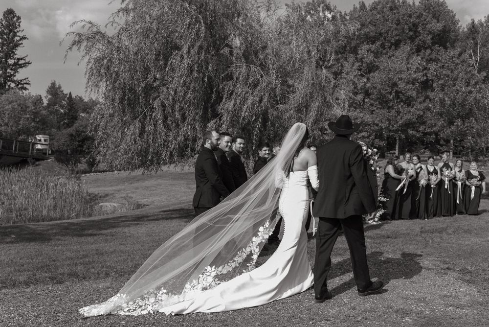 A wedding ceremony in a garden setting with a long flowing veil trailing behind the couple in black and white.