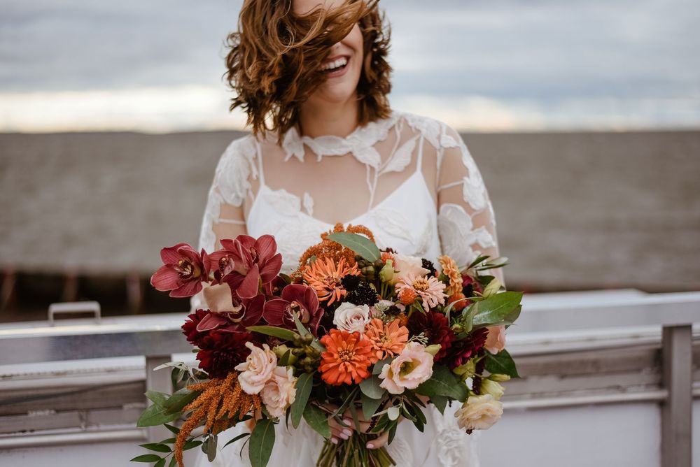 A delicate peach and burgundy bridal bouquet with roses and foliage held in front of a stone wall backdrop.