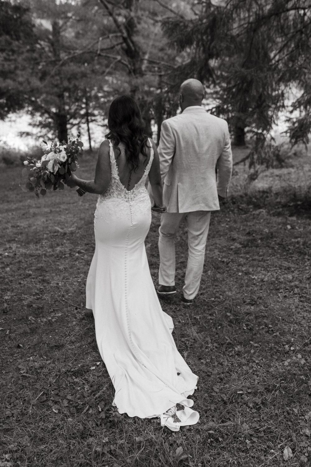 Black and white photo of a bride in a fitted lace gown walking behind a groom in a light suit outdoors in nature.
