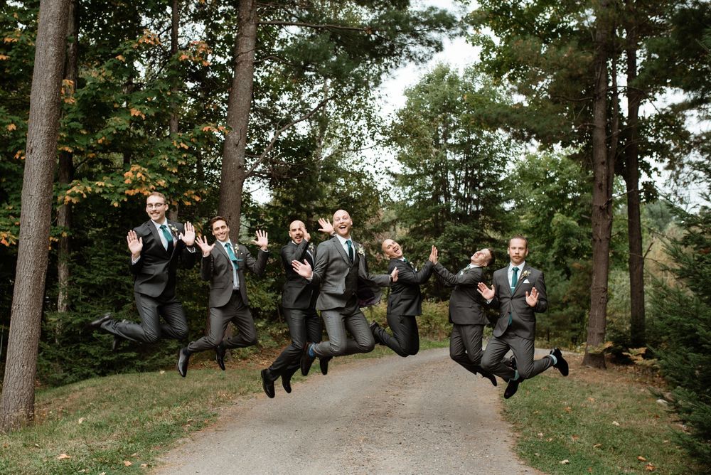 A group in matching grey suits jumps joyfully in sync on a wooded path during a wedding celebration.