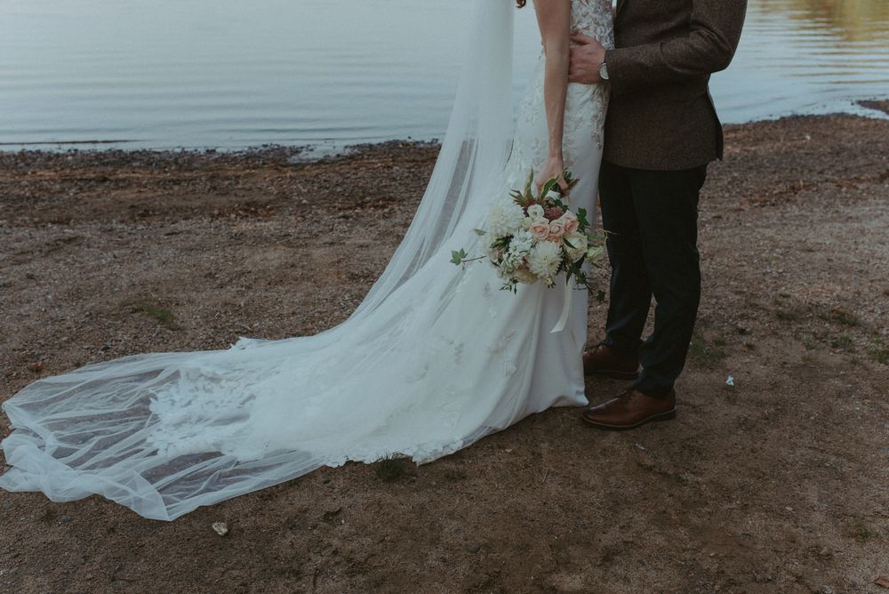 A wedding couple stands at the water's edge on a beach during sunset while their bridal gown train flows in the breeze.