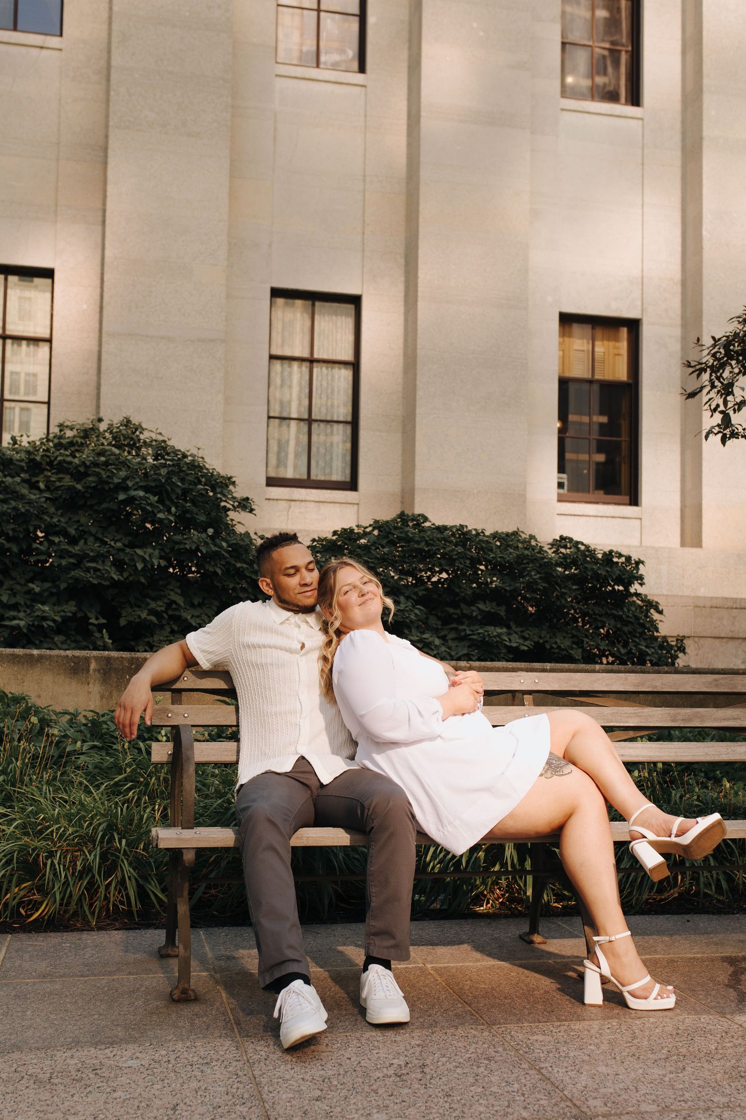 Engaged couple sitting closely on a bench outside the Ohio Statehouse in warm morning sunlight.