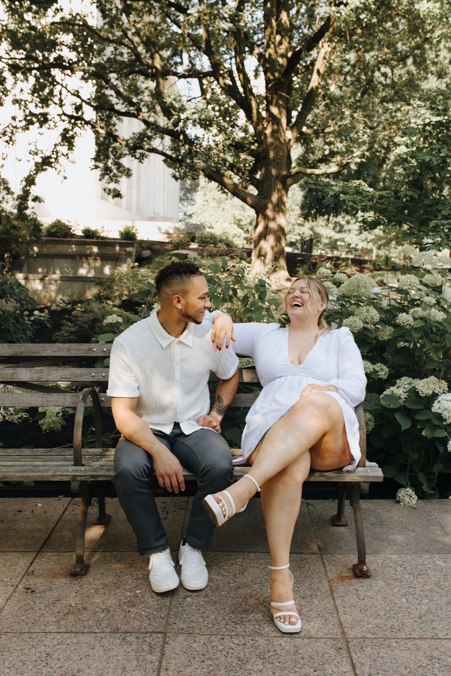 Natural light engagement photo of couple sitting and laughing on a bench outside the Ohio Statehouse.