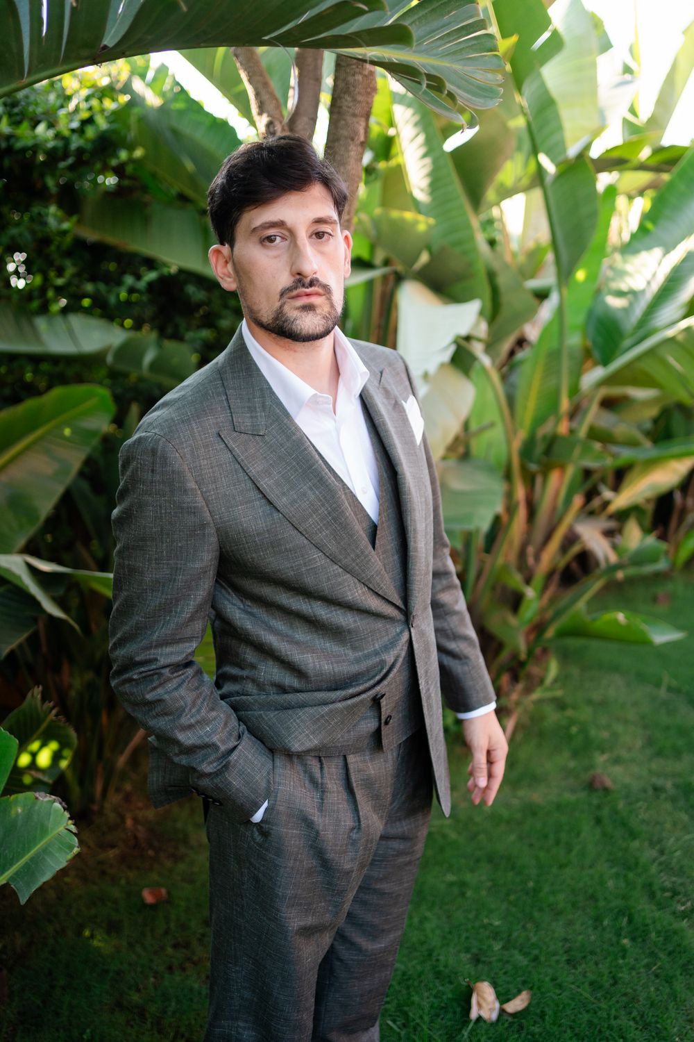 A person in a grey suit poses among tropical plants and greenery in an outdoor setting.