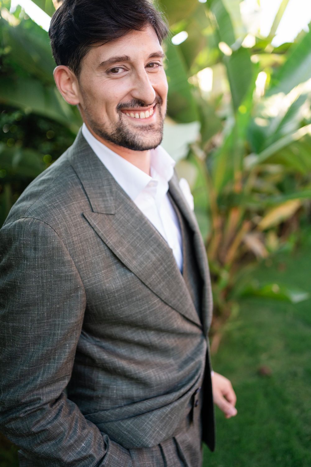 A person in a grey suit smiles while posing in a garden setting with tropical plants.