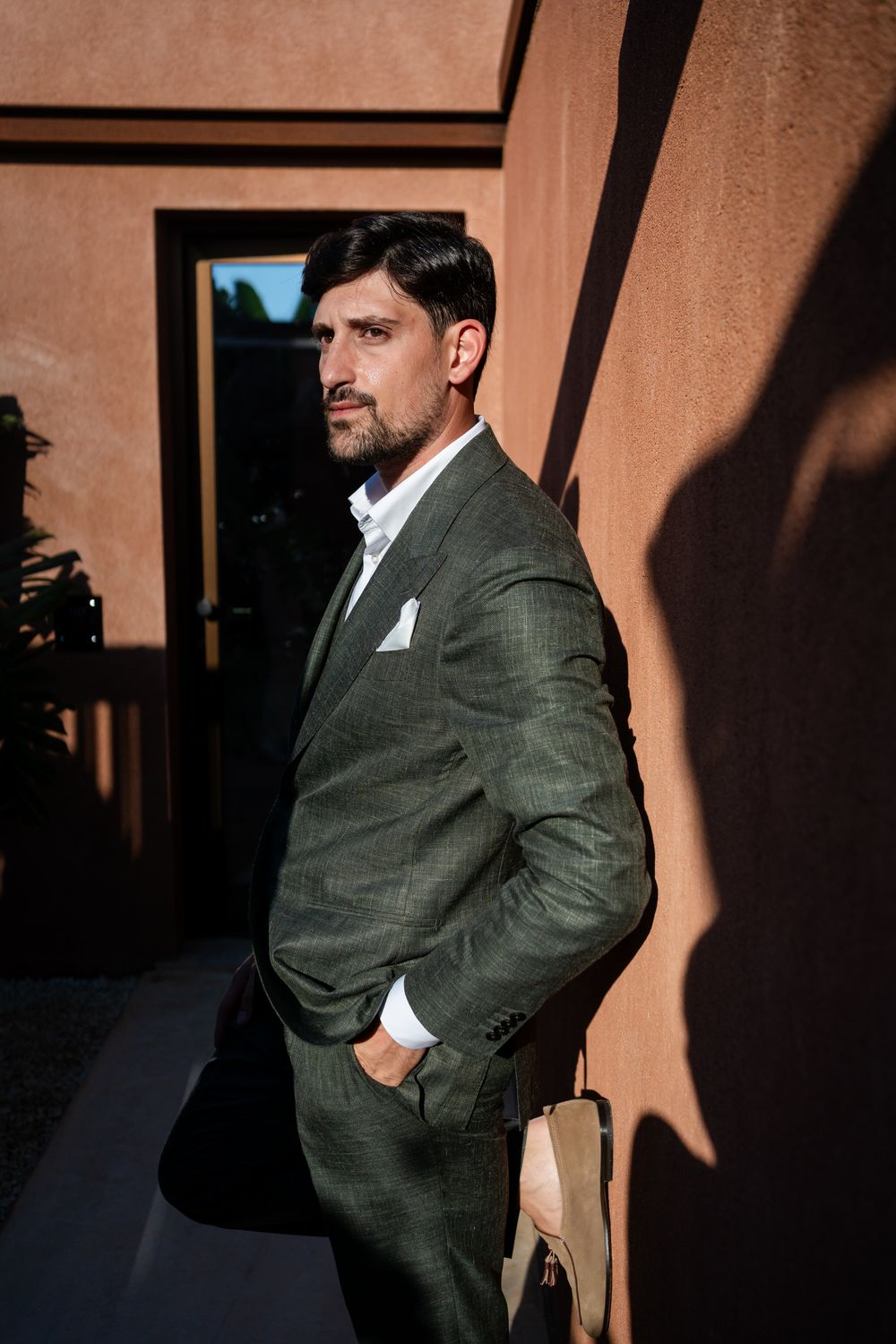 A man in a grey suit and white pocket square poses against a terracotta wall.