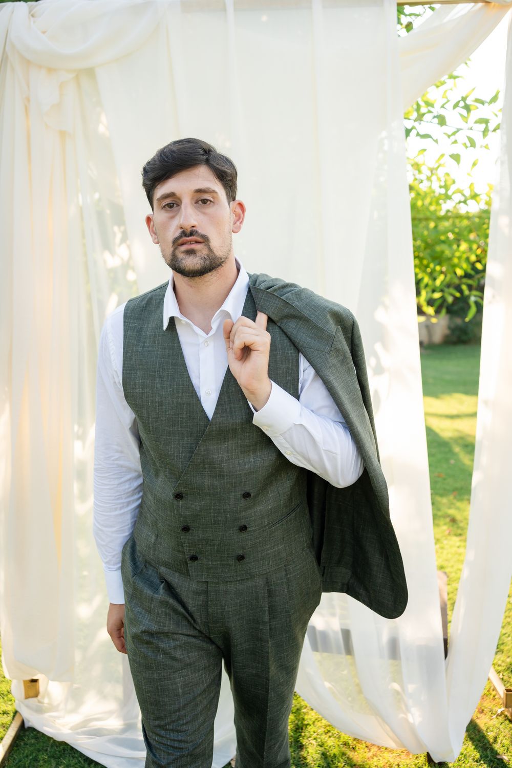A person in a grey double-breasted vest and white shirt stands against white drapery.