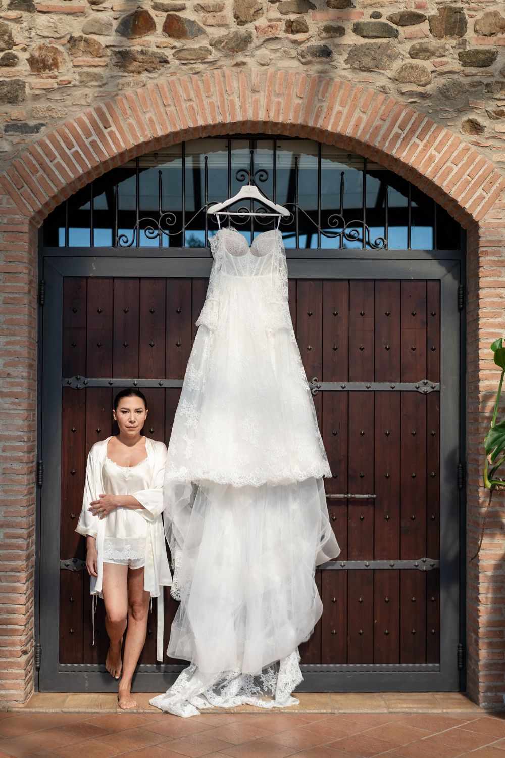 White wedding dress hangs displayed against rustic stone wall with wooden arched doors.