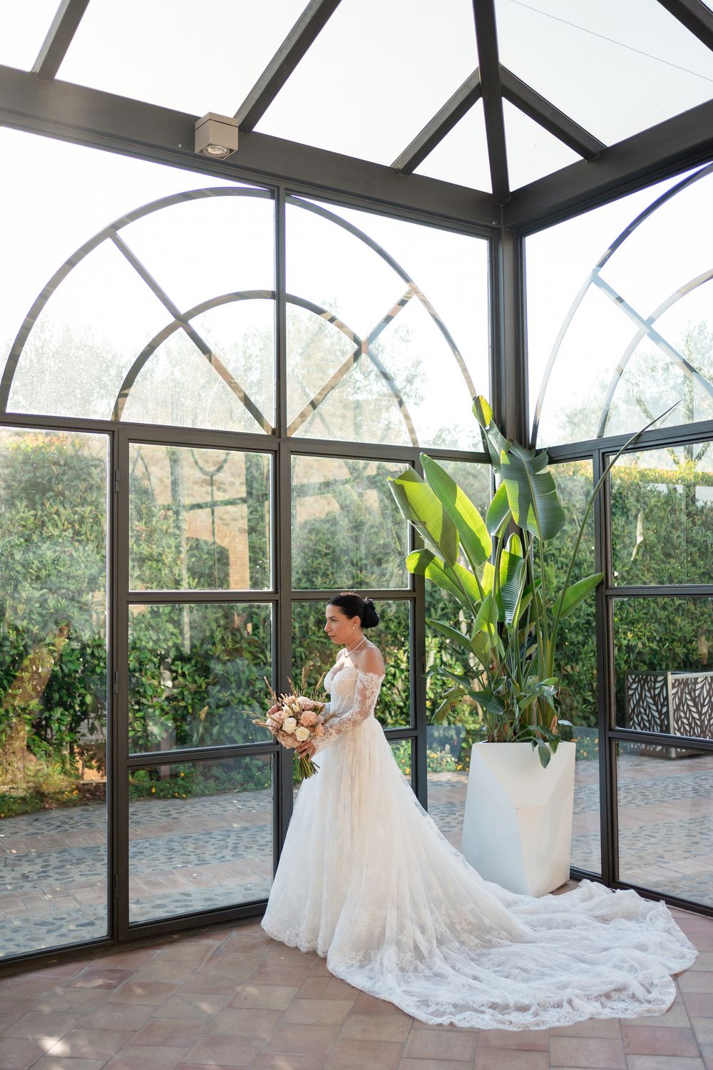 Stunning bride in lace gown with long train standing in glass conservatory.