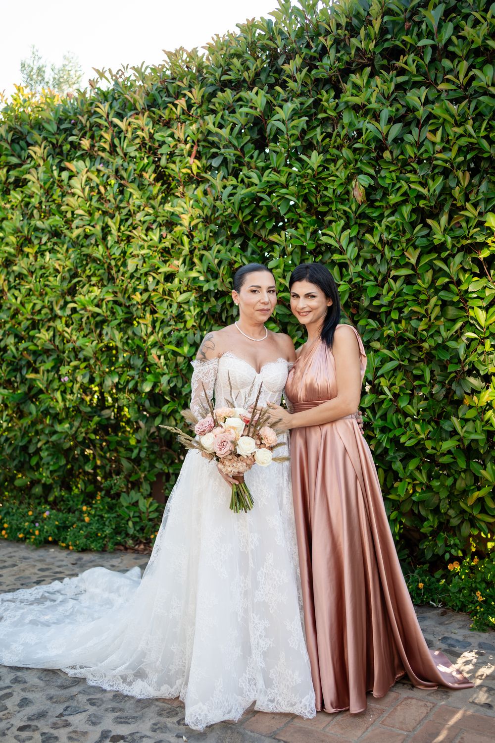 Wedding party poses in front of lush green hedge with bride in white lace gown and attendant in rose gold dress.