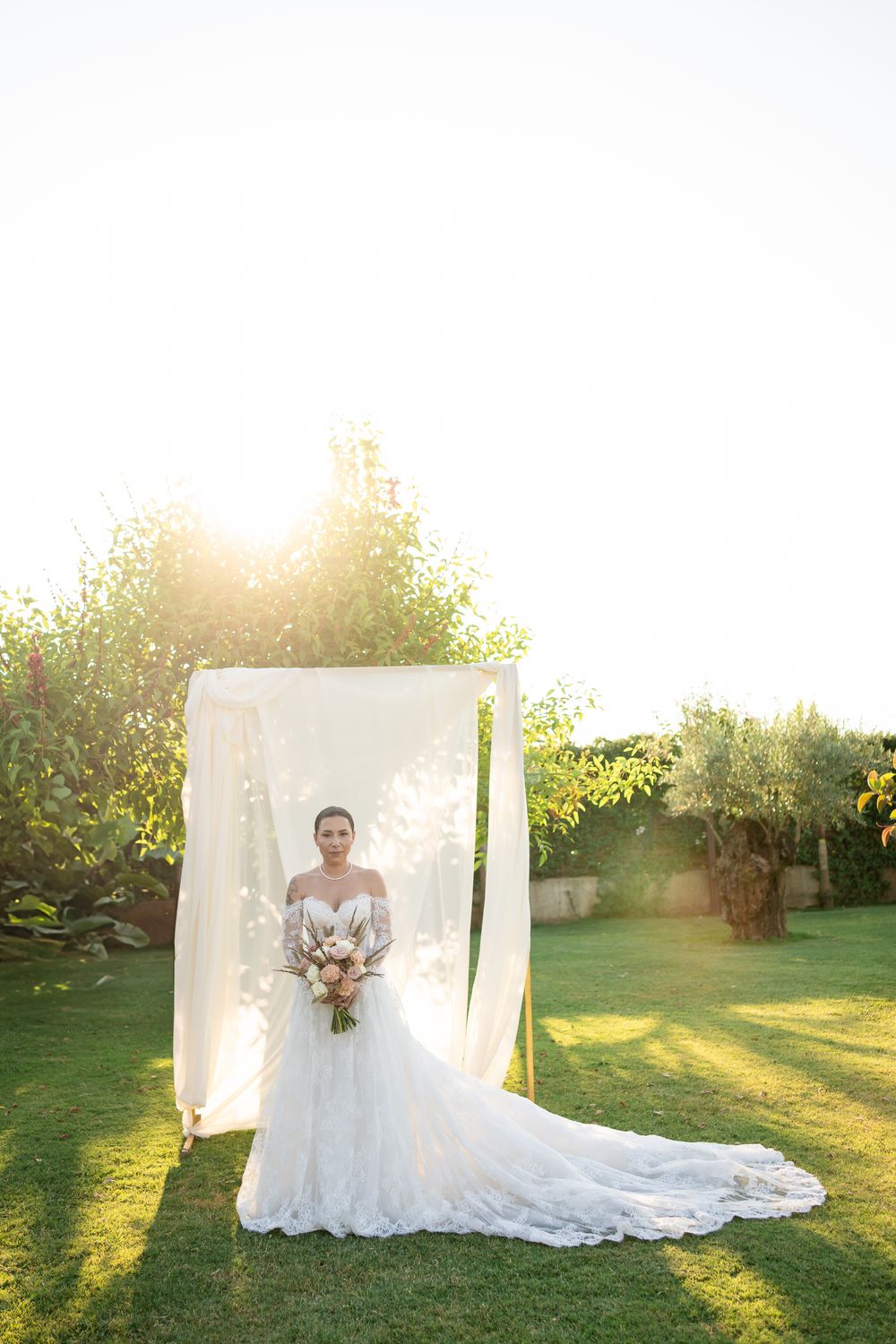 Bride in white lace gown stands by draped backdrop in sunlit garden setting.