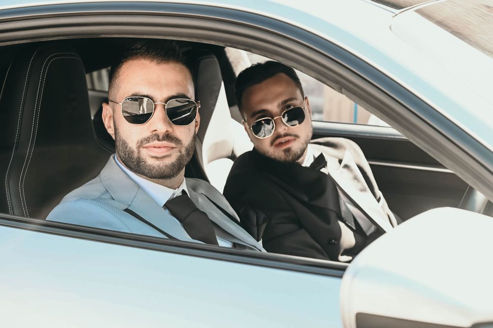 Two well-dressed men wearing sunglasses sit in the front seats of a luxury car.