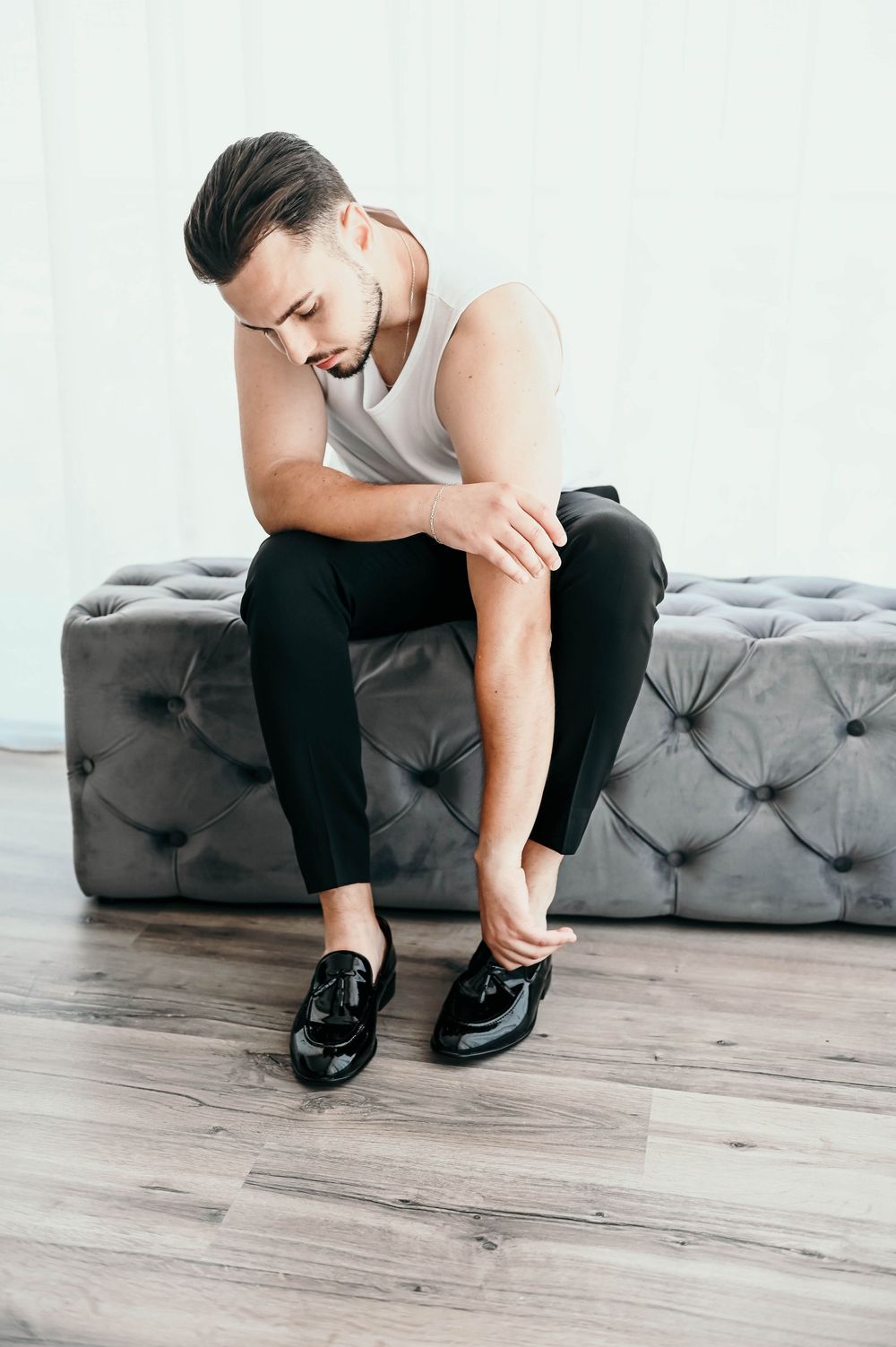 Person sitting on a grey tufted ottoman wearing black pants and white tank top while putting on dress shoes.