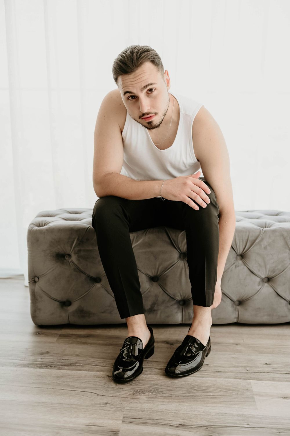 A person in a white tank top and black pants sits thoughtfully on a gray tufted couch against a white wall.