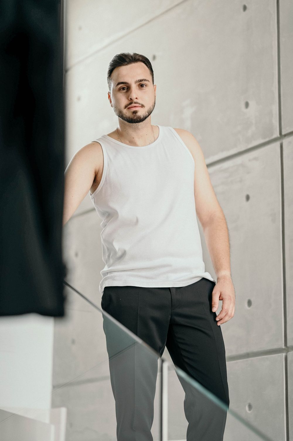 Fitness model wearing white tank top and black pants posing against concrete wall in modern interior setting.
