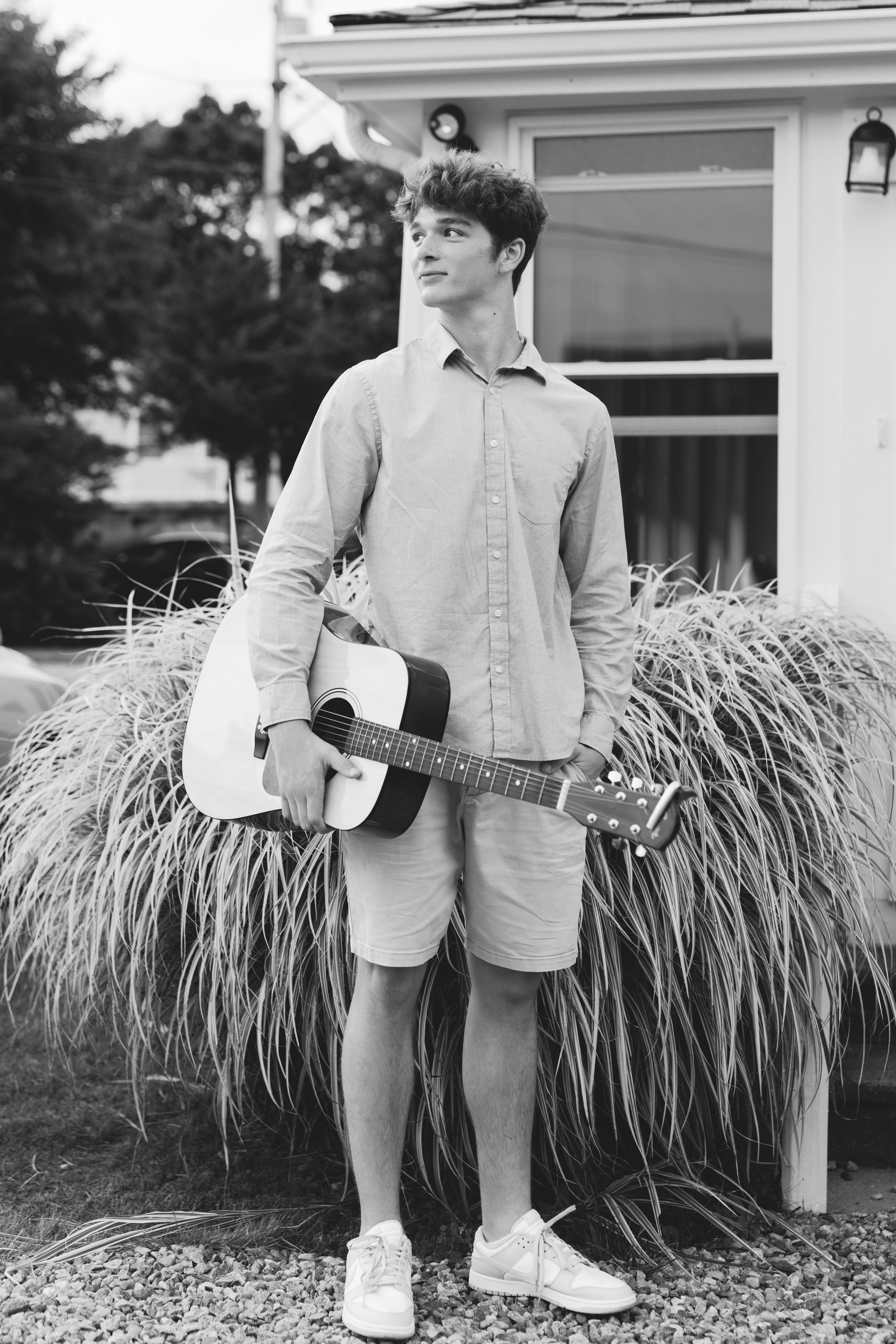 Black and white photo of a young person in casual attire holding an acoustic guitar while standing by ornamental grass.