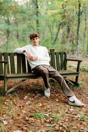 A person in white sits on a wooden park bench surrounded by fallen autumn leaves in a wooded setting.
