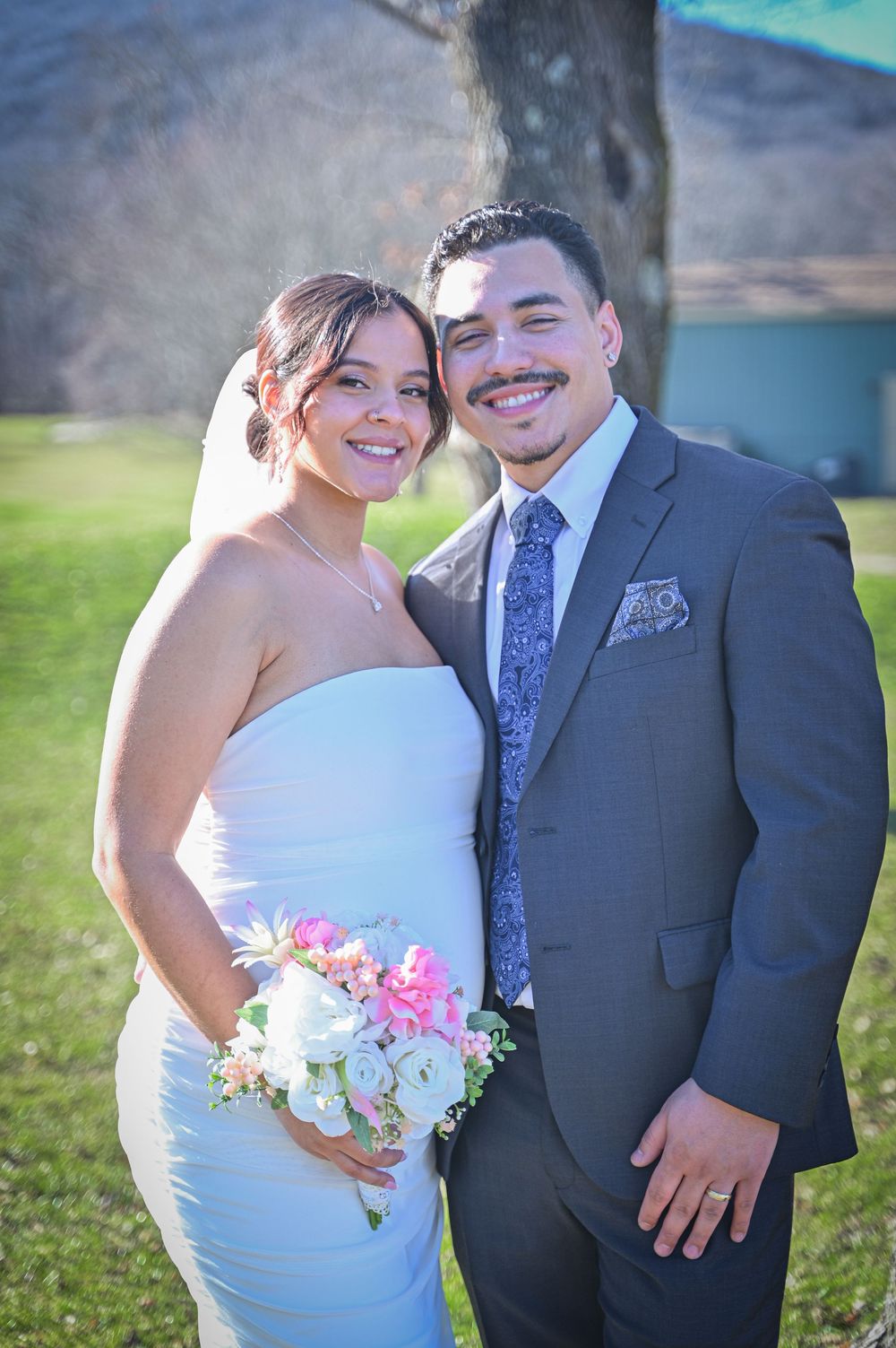 A couple in wedding attire stands together in a sunny outdoor setting with colorful floral bouquet.