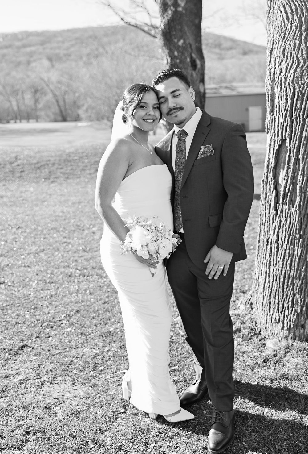A black and white outdoor portrait of a wedding couple standing by trees with mountain views in the background.