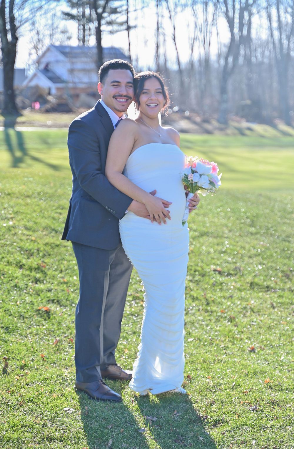 A couple shares a joyful embrace on their wedding day in a sunlit grassy field with trees in the background.