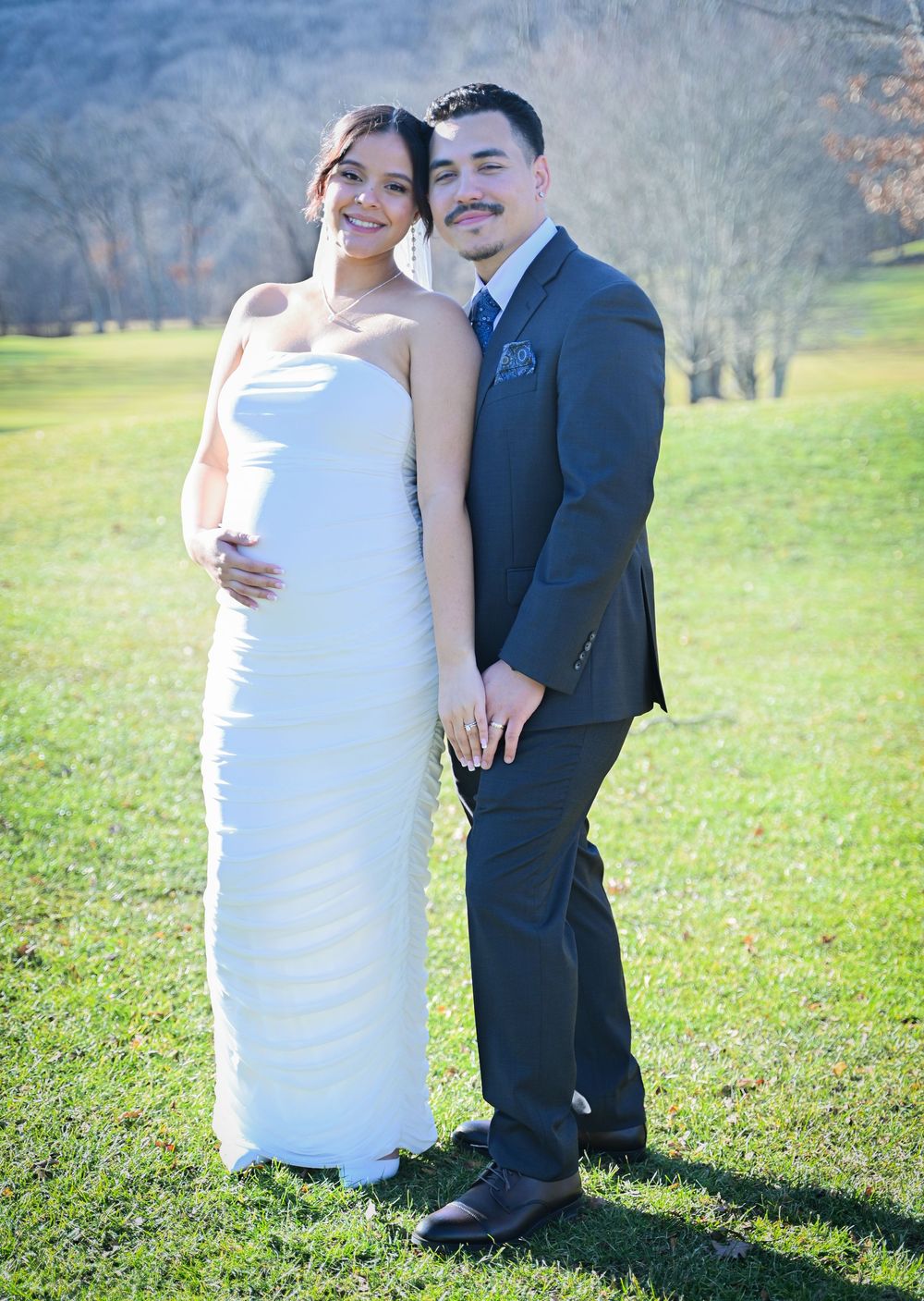 A couple in wedding attire stands together outdoors on grass with trees in the background.