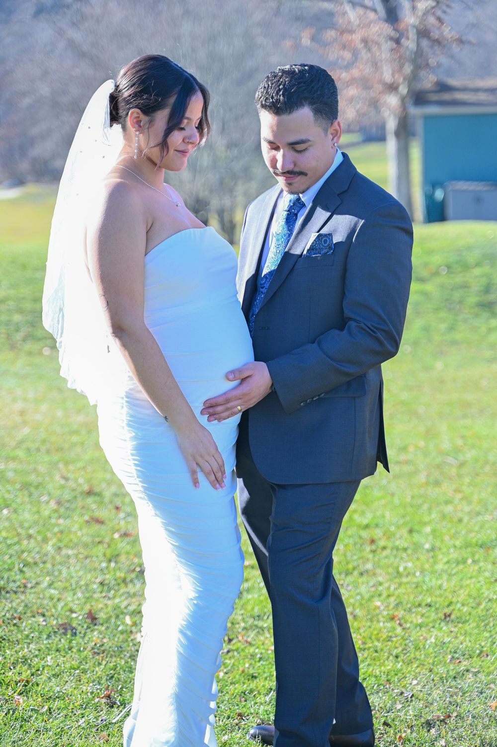 Pregnant woman in a white wedding dress standing with partner in navy suit during outdoor maternity photoshoot.