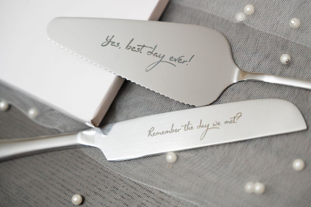 Two wedding cake knives with romantic engraved messages on their silver blades lying on a gray surface.