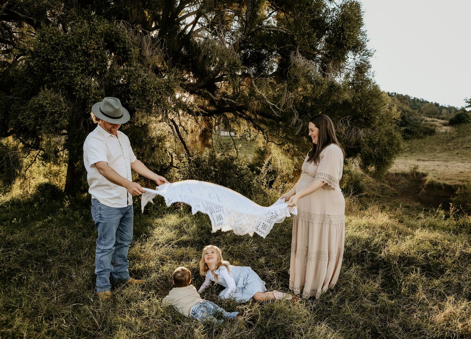 Family members in neutral colored clothing pose together on a grassy hillside during golden hour photography session.