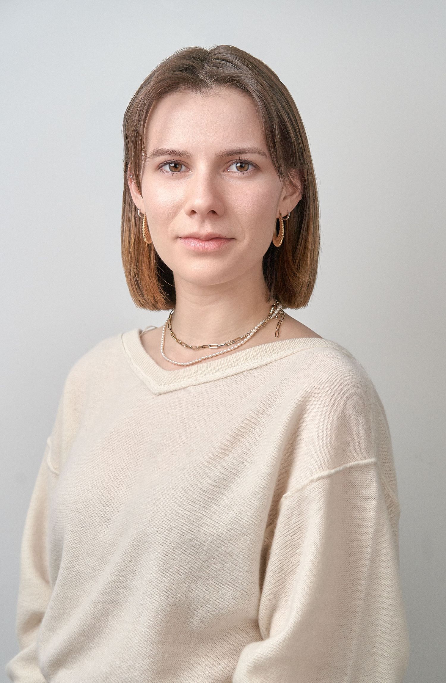 Young woman with bob haircut in beige sweater and gold necklace in professional headshot.