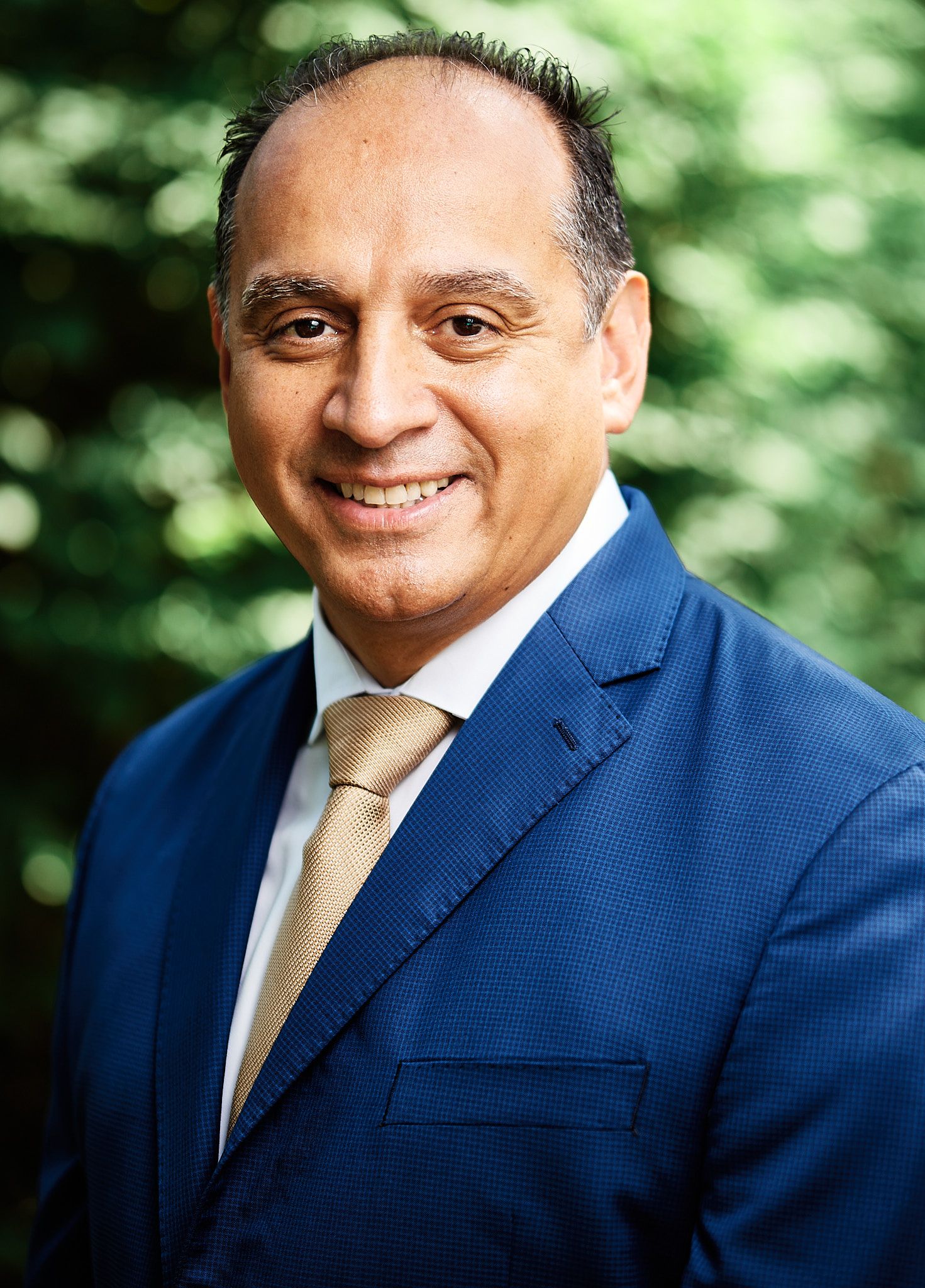 Smiling professional man in blue suit and gold tie posing outdoors for business headshot.