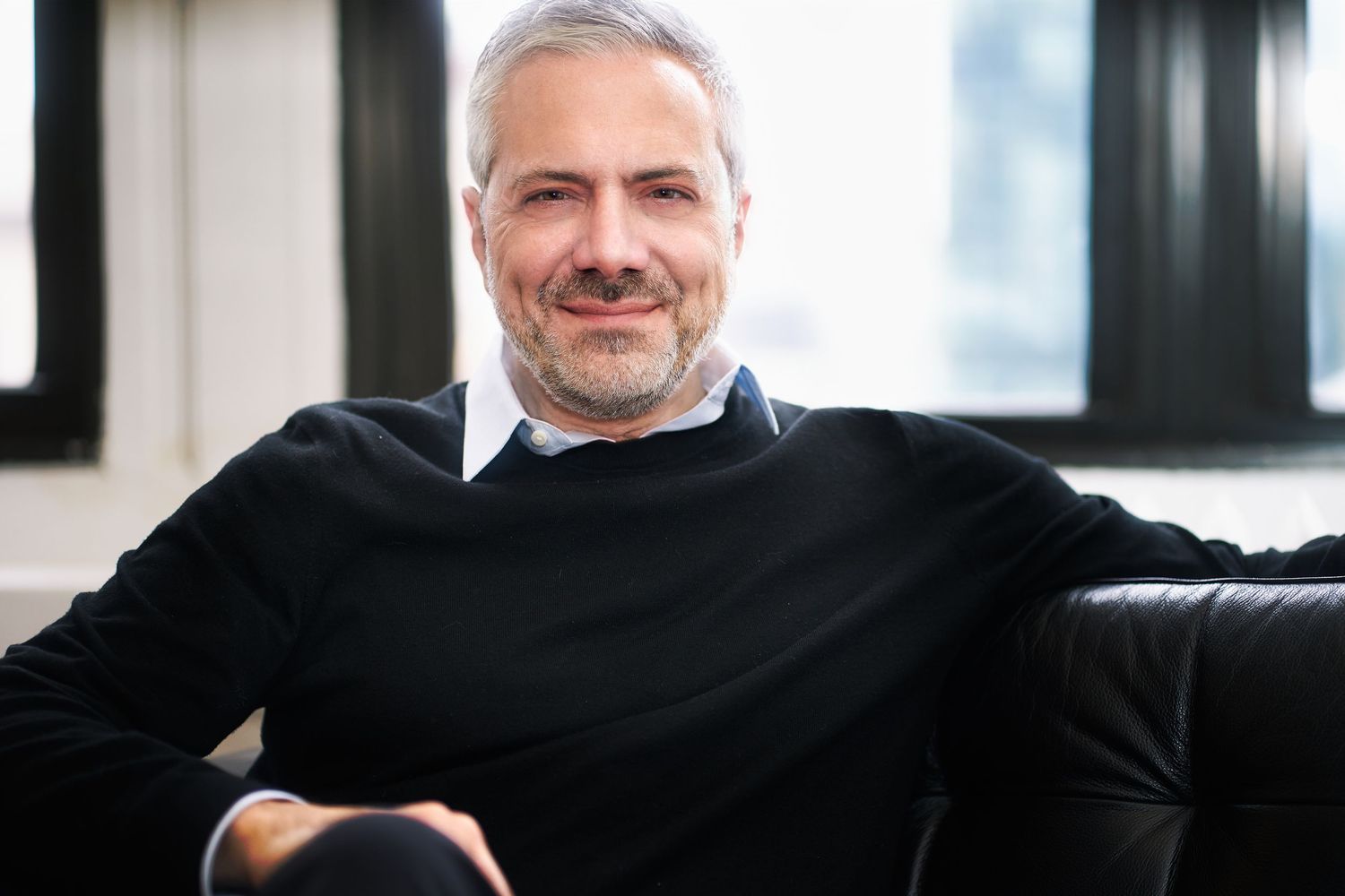 Smiling middle-aged man with gray hair and beard wearing a black sweater, seated on a leather sofa.