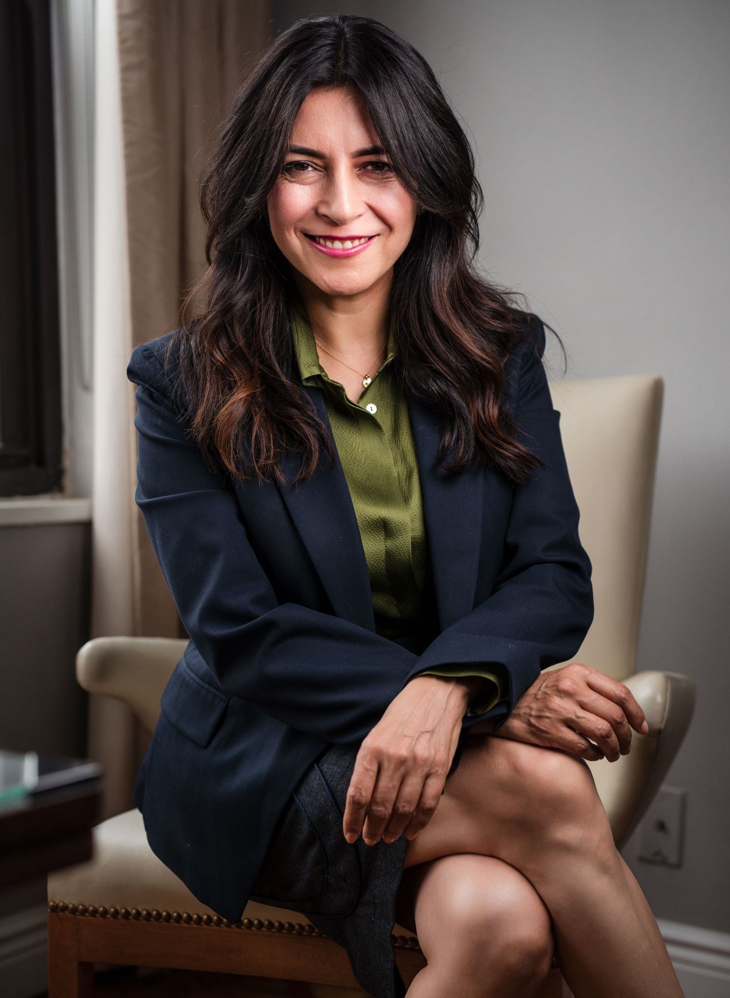 Young professional woman in a navy blazer and green shirt seated in a chair with a warm smile.