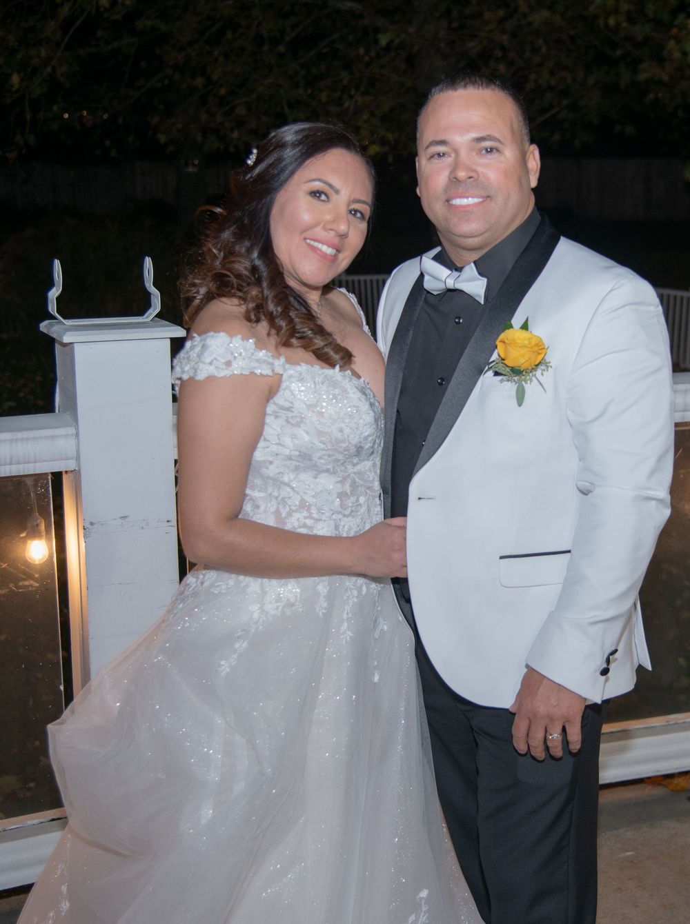 A happy couple smiling together at their wedding reception, wearing white formal attire with a boutonniere on the groom.