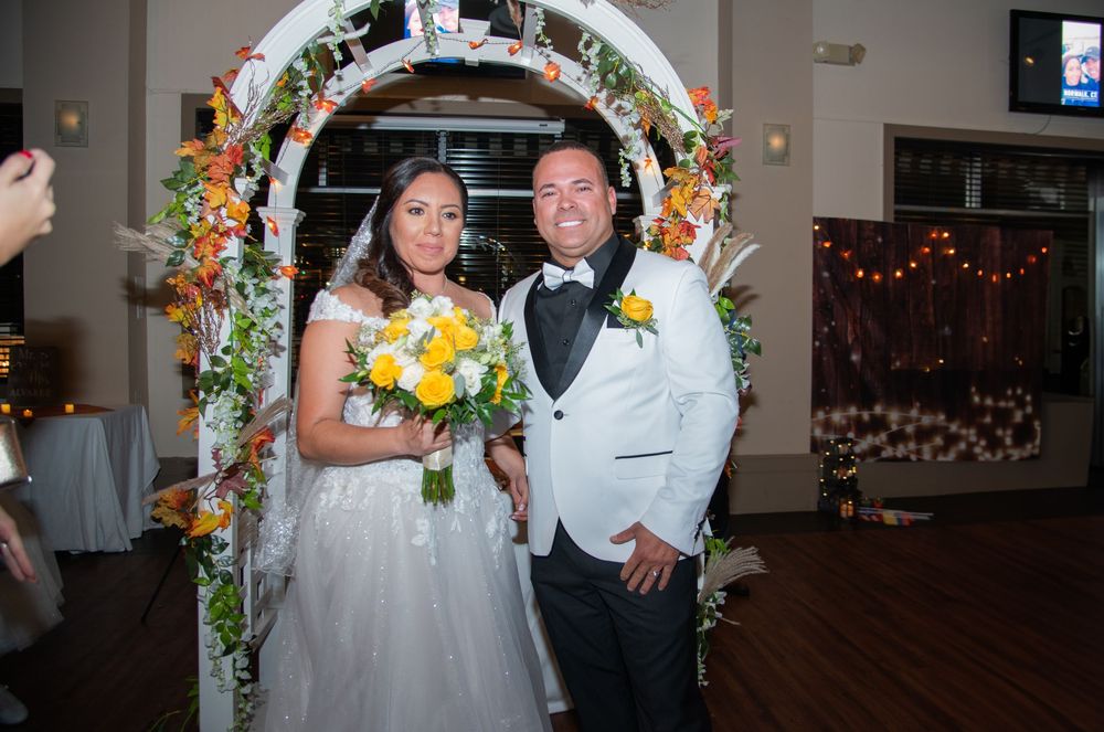 Newlyweds stand under floral arch decorated with orange and yellow flowers at indoor reception venue.