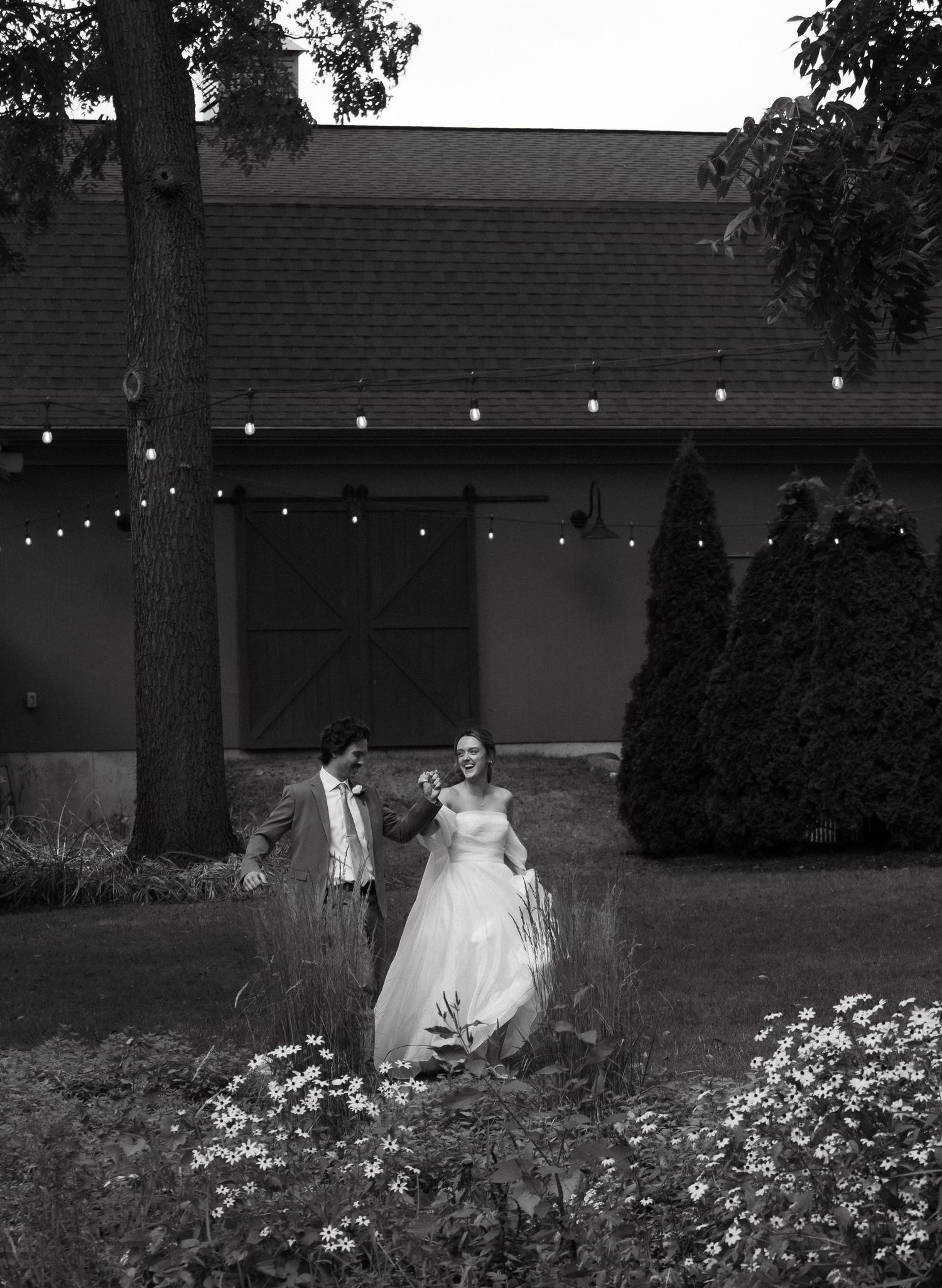 Black and white photo of newlyweds walking through a garden with string lights overhead.