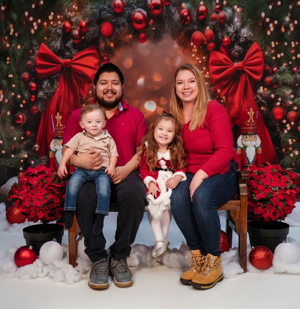 Family wearing red and denim sits on wooden bench against Christmas backdrop with ornaments and bows.
